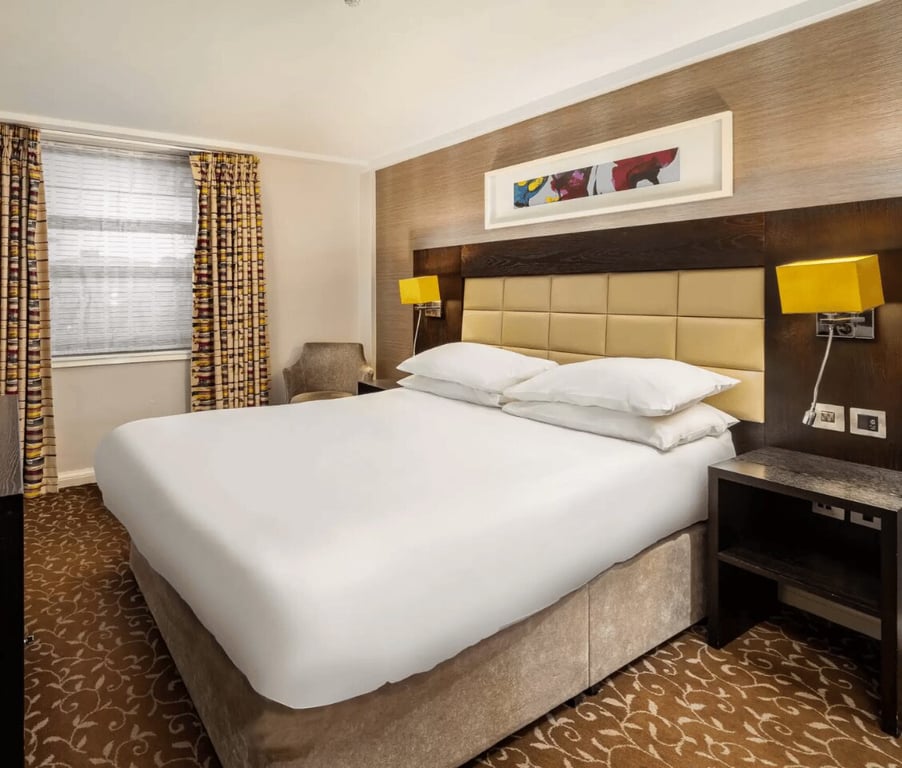 Hotel room at The Lensbury with a classic double bed, cushioned headboard, mustard lamps, patterned carpet, and curtains.