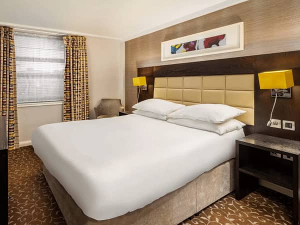 Hotel room at The Lensbury with a classic double bed, cushioned headboard, mustard lamps, patterned carpet, and curtains.