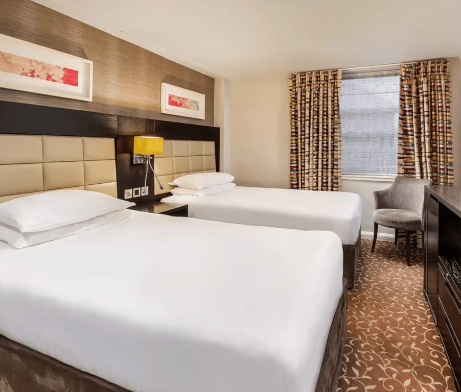 The Lensbury hotel classic twin room with two single beds, a yellow bedside lamp, modern art on the walls, and a window with patterned curtains.