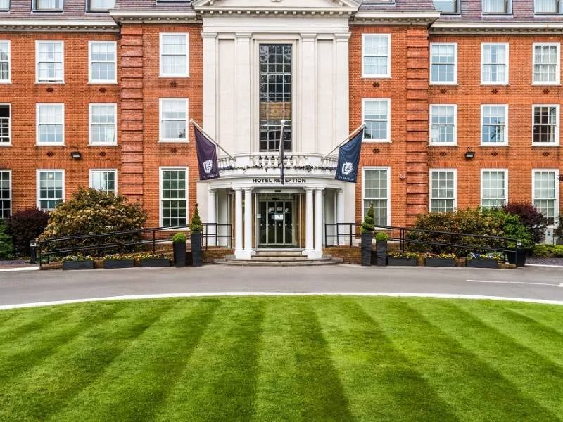 Front view of The Lensbury hotel entrance, showcasing its red-brick facade, orderly lawns, and flags above the revolving door entrance.
