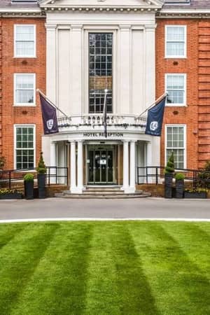 Front view of The Lensbury hotel entrance, showcasing its red-brick facade, orderly lawns, and flags above the revolving door entrance.