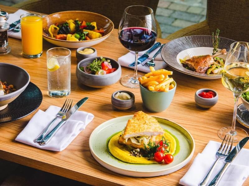 Dining table at The Lensbury featuring gourmet dishes such as roasted chicken, pasta, salad, and wine.