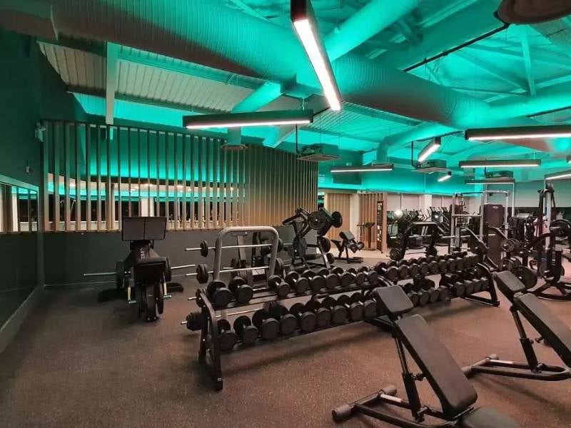 Modern gym at The Lensbury with free weights, benches, and cardio machines under green ambient lighting.