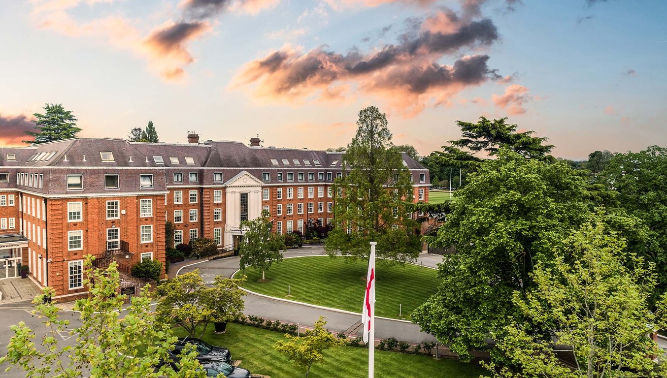 Aerial view of The Lensbury Hotel, showing a red-brick building with a circular drive and lush trees, set under a sunset sky.
