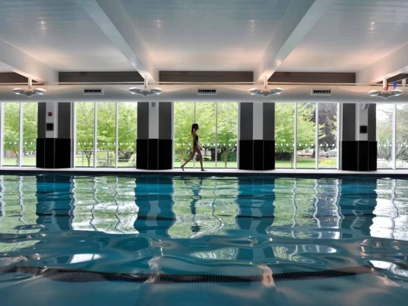 Indoor pool at The Lensbury with a person walking alongside and large windows displaying green trees outside.