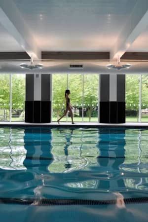 Indoor pool at The Lensbury with a person walking alongside and large windows displaying green trees outside.