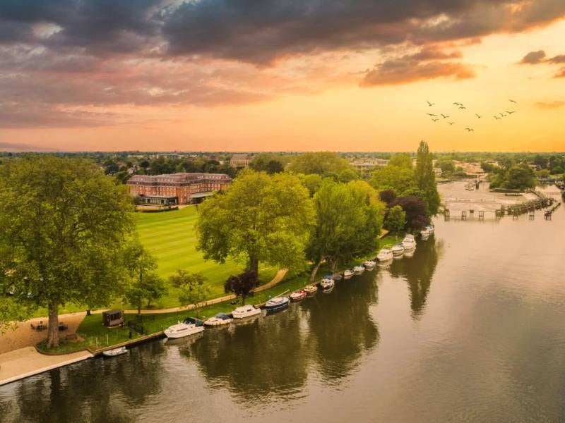 Aerial view of The Lensbury resort on the River Thames, with boats, lush greenery, and a sunset sky.