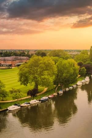 Aerial view of The Lensbury resort on the River Thames, with boats, lush greenery, and a sunset sky.