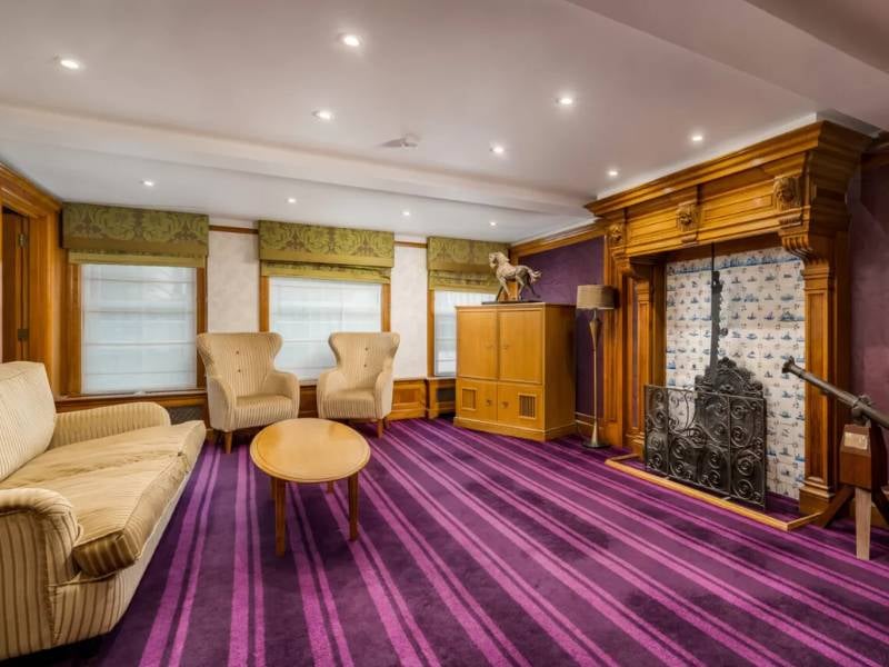 Luxurious room at The Lensbury with beige seating, wooden table, ornate fireplace, purple striped carpet, and large windows with green valances.