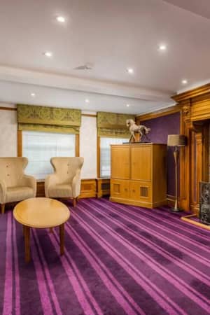 Luxurious room at The Lensbury with beige seating, wooden table, ornate fireplace, purple striped carpet, and large windows with green valances.