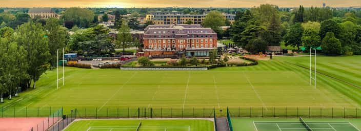 Aerial view of The Lensbury sports facilities with tennis courts, rugby fields, and a clubhouse set against a sunset backdrop.