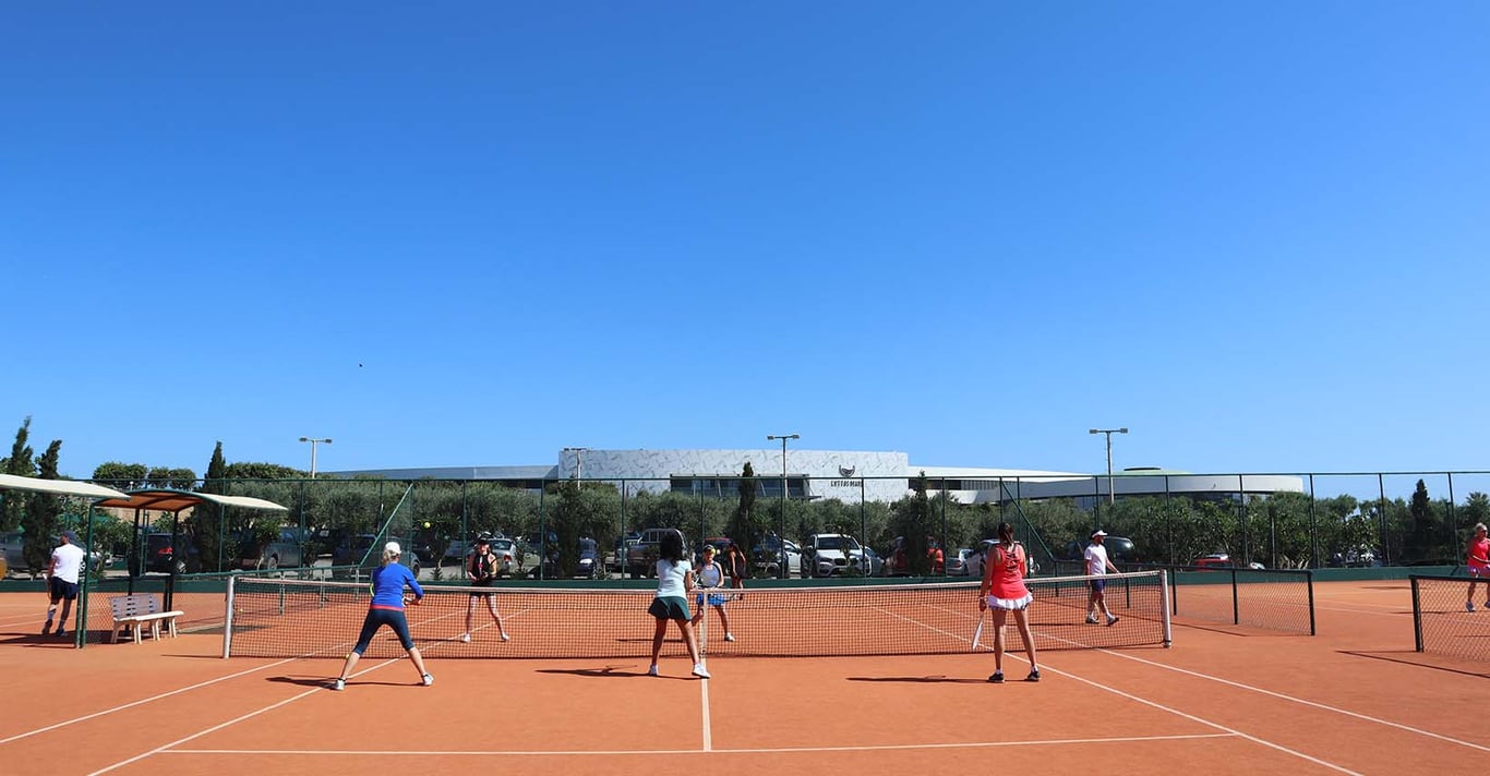 Group playing doubles tennis on an outdoor clay court, marking the start of the 2024 tennis holiday season with clear skies and modern facilities in the background.