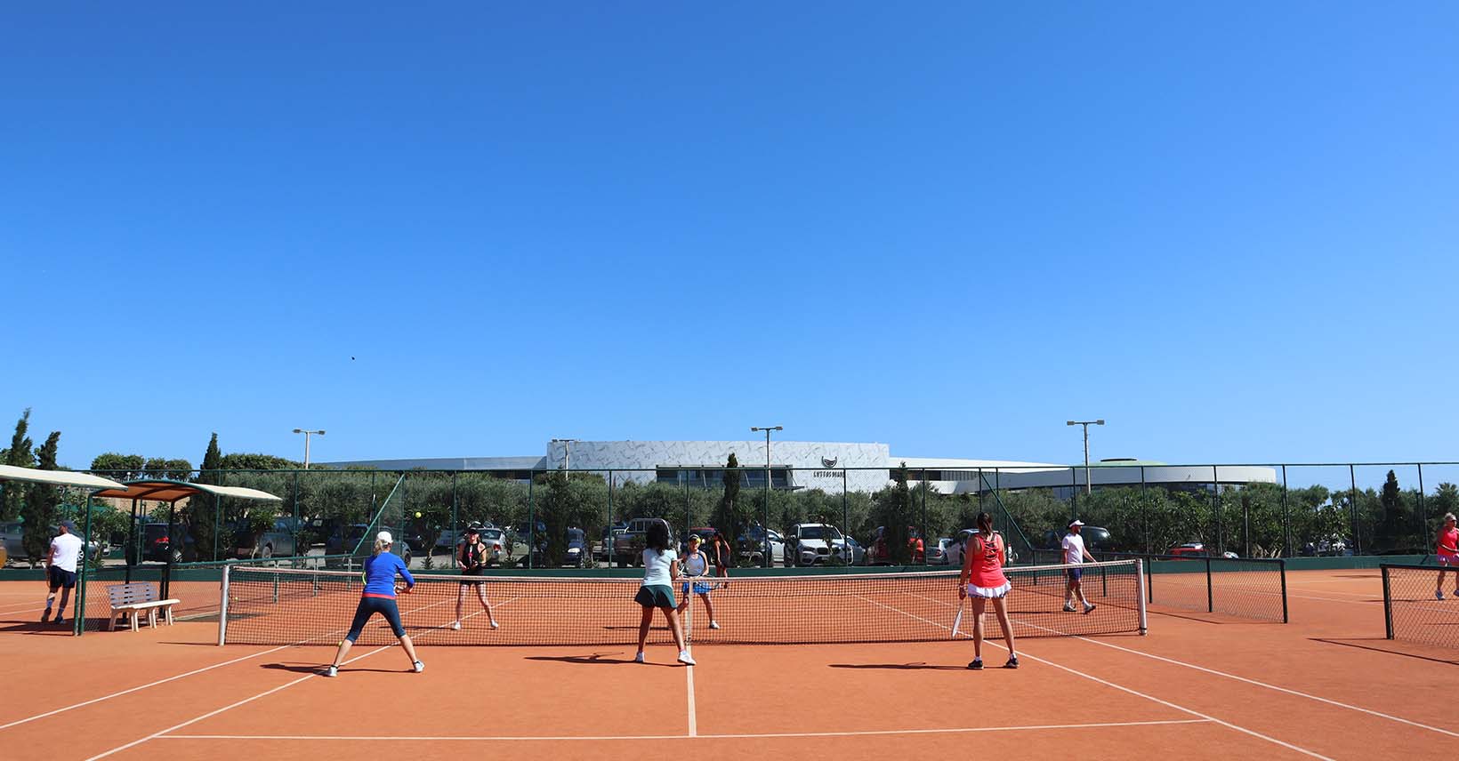 Group playing doubles tennis on an outdoor clay court, marking the start of the 2024 tennis holiday season with clear skies and modern facilities in the background.