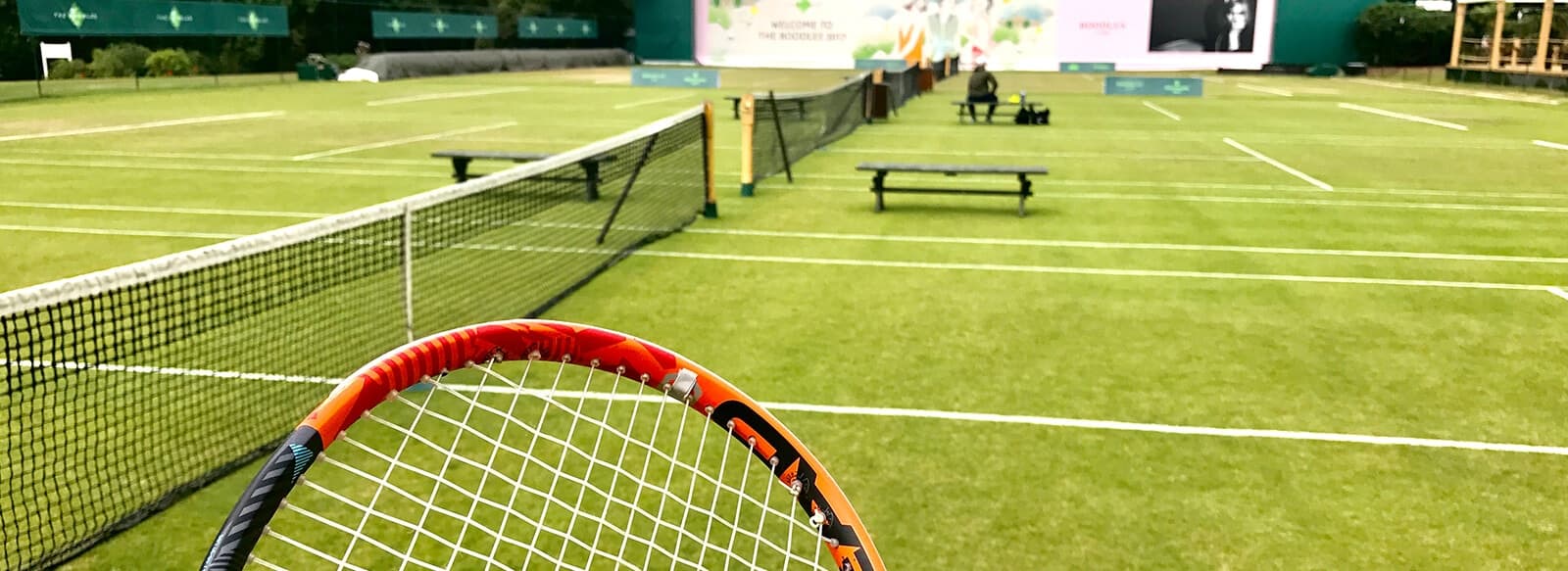 Grass tennis court with white lines, featuring a tennis racket in the foreground and a large event screen in the background.