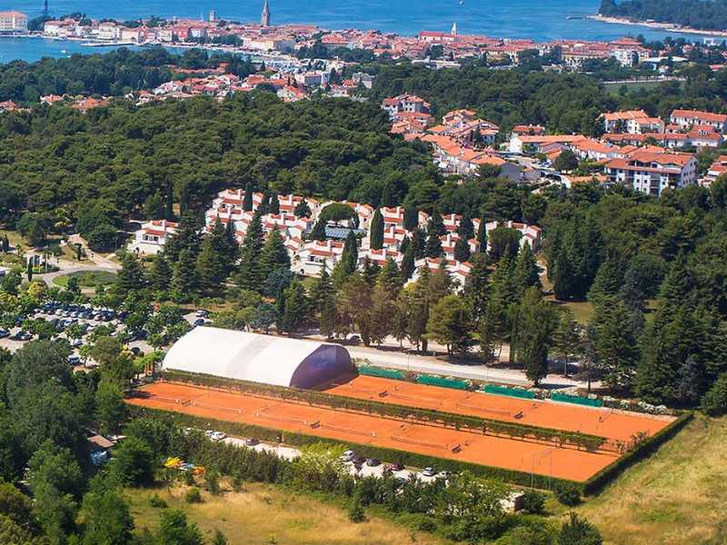 Aerial view of Valamar clay tennis courts surrounded by trees and residential buildings, with the Adriatic Sea and a coastal town in the background.