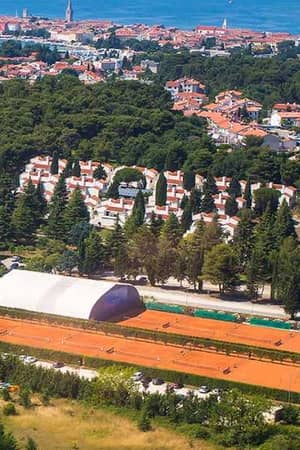 Aerial view of Valamar clay tennis courts surrounded by trees and residential buildings, with the Adriatic Sea and a coastal town in the background.