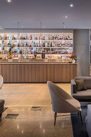 Valamar Gallery Lounge with a modern bar, illuminated bottle shelves, grey armchairs, and sofas, attended by two staff members.