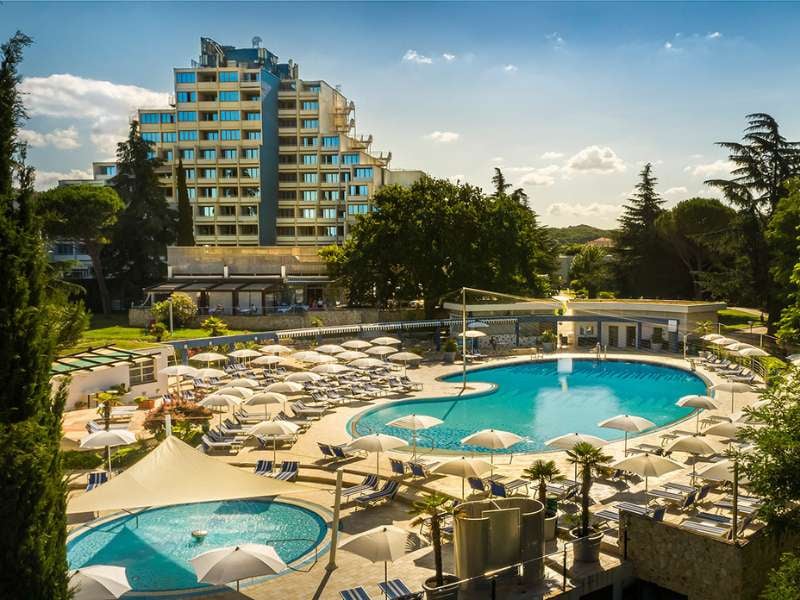 Valamar hotel exterior with a wide view of the pool lounge area, sun loungers and parasols, surrounded by greenery.
