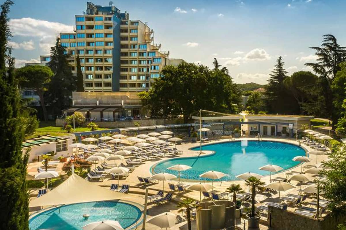 Valamar hotel exterior with a wide view of the pool lounge area, sun loungers and parasols, surrounded by greenery.