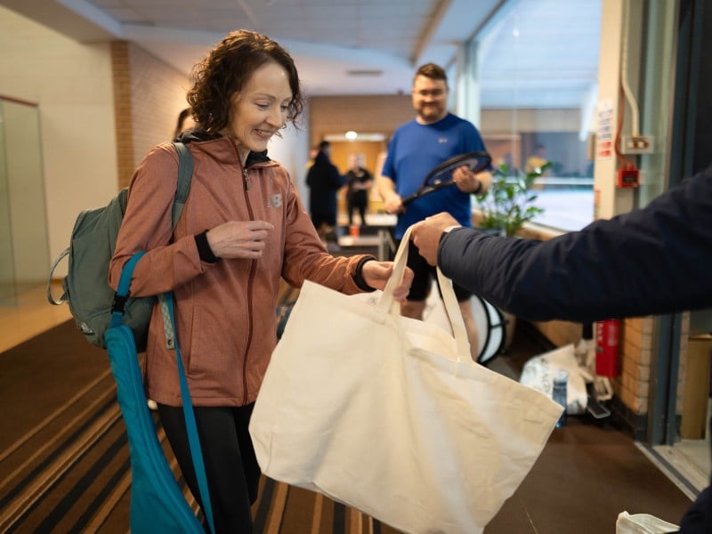 Woman in sporty outfit receiving a tote bag from a host at Virgin Active Chiswick gym, with other people and staff in the background.