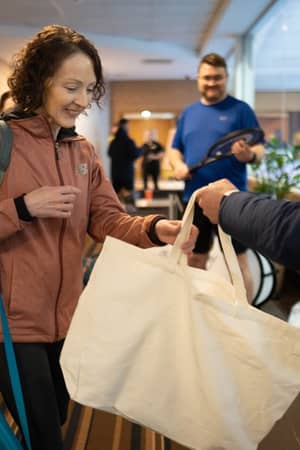 Woman in sporty outfit receiving a tote bag from a host at Virgin Active Chiswick gym, with other people and staff in the background.