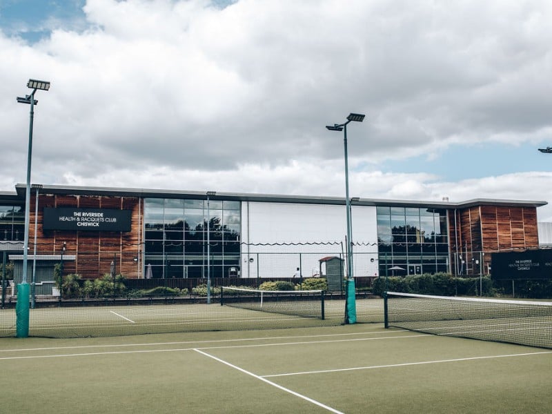 Exterior of Virgin Active Chiswick showing a modern building with glass and wood design, and a tennis court in the foreground.
