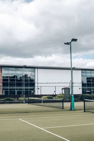 Exterior of Virgin Active Chiswick showing a modern building with glass and wood design, and a tennis court in the foreground.