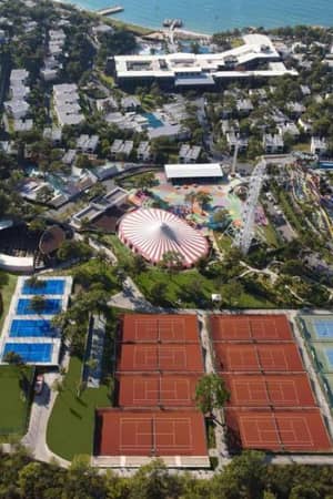 Aerial view of a sports complex with red clay tennis courts and blue hard courts near a coastline and theme park.