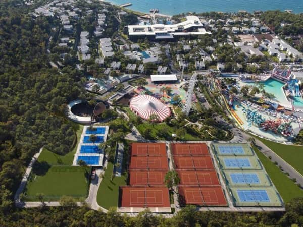 Aerial view of a sports complex with red clay tennis courts and blue hard courts near a coastline and theme park.