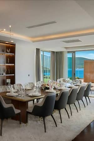 Luxurious dining room with a table set for dining, grey chairs, a white rug, and large windows overlooking the ocean.