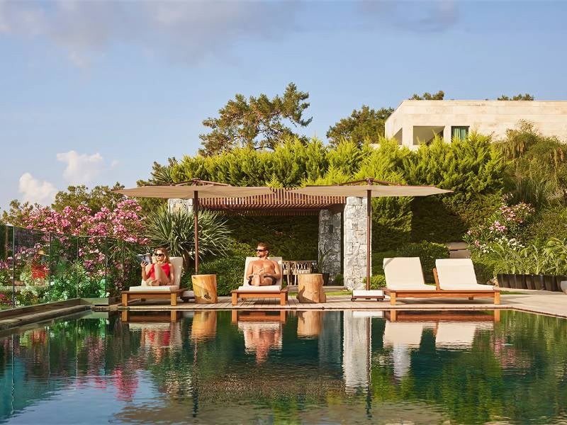 Outdoor poolside view with two people on sun loungers, shaded by parasols, surrounded by greenery and flowering plants.