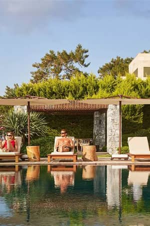 Outdoor poolside view with two people on sun loungers, shaded by parasols, surrounded by greenery and flowering plants.