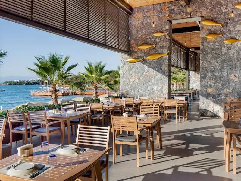 Vogue Gallery restaurant's dining area with wooden furniture, stone walls, golden fish decor, and a view of palm trees and the ocean.