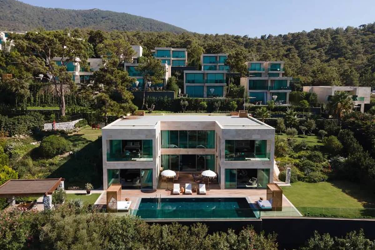 Luxurious modern villa with large glass windows and a swimming pool, set in a green hillside with surrounding upscale residences.