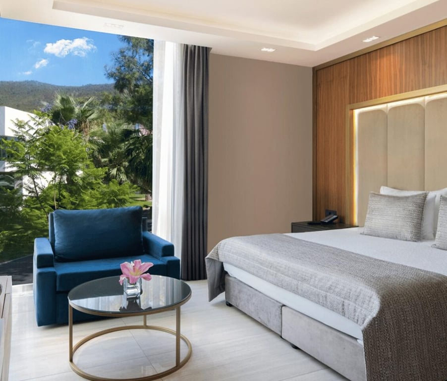 Modern hotel room in Vogue Garden Superior Annex with a king-size bed, blue armchair, and a view of greenery through a large window.
