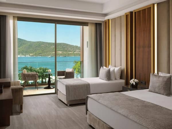 Vogue Superior Room with two twin beds, offering a sea view through floor-to-ceiling windows and a balcony, decorated in soft neutrals with wood accents.