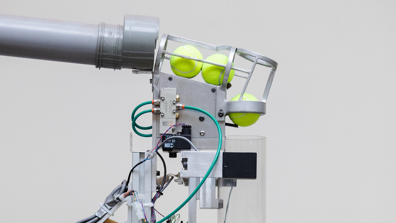 Tennis ball launcher with a metal cage holding tennis balls and connected to pipes and wires for advanced practice sessions.