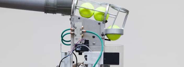 Tennis ball launcher with a metal cage holding tennis balls and connected to pipes and wires for advanced practice sessions.