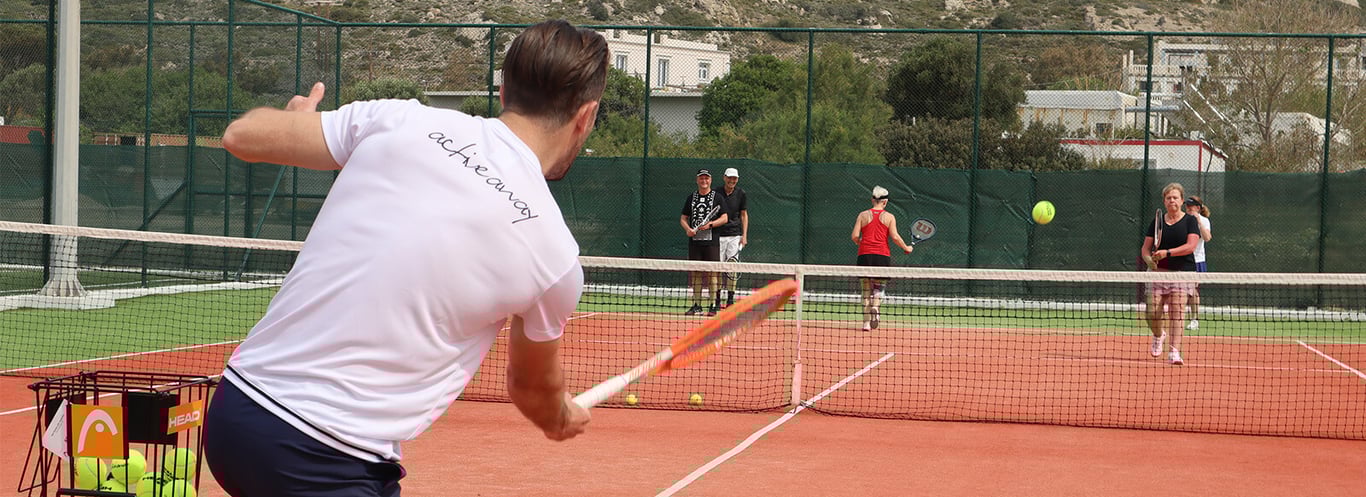 Tennis coach performing a backhand swing during a group session on an outdoor court with three students in the background, part of Active Away's 2024 programme.