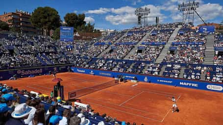 Barcelona Open tennis match in progress, showing players on clay courts and a large audience in the stands under a sunny sky.