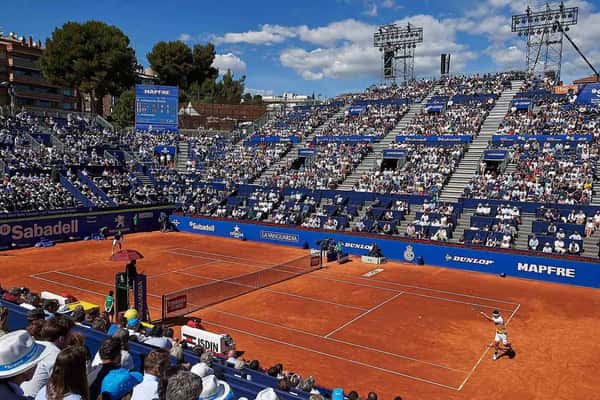 Barcelona Open tennis match in progress, showing players on clay courts and a large audience in the stands under a sunny sky.