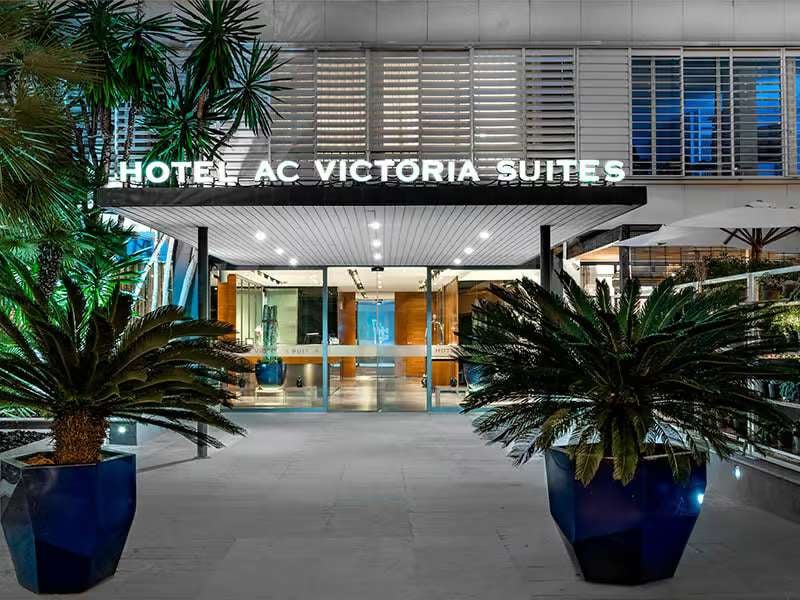 Entrance of Hotel AC Victoria Suites in Barcelona, with glass doors, metal design, and potted palms.