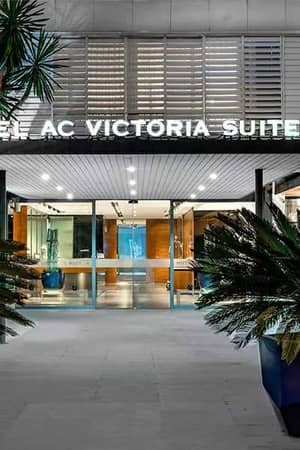 Entrance of Hotel AC Victoria Suites in Barcelona, with glass doors, metal design, and potted palms.