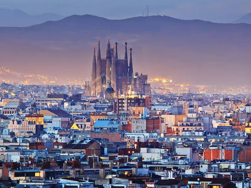 Nighttime image of Barcelona's cityscape featuring the illuminated Sagrada Família basilica, set against a mountainous backdrop.