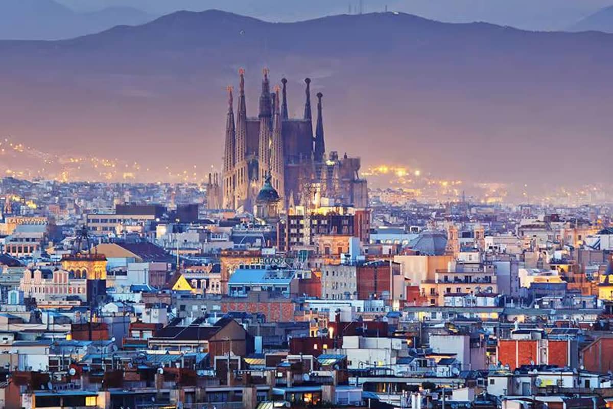 Nighttime image of Barcelona's cityscape featuring the illuminated Sagrada Família basilica, set against a mountainous backdrop.