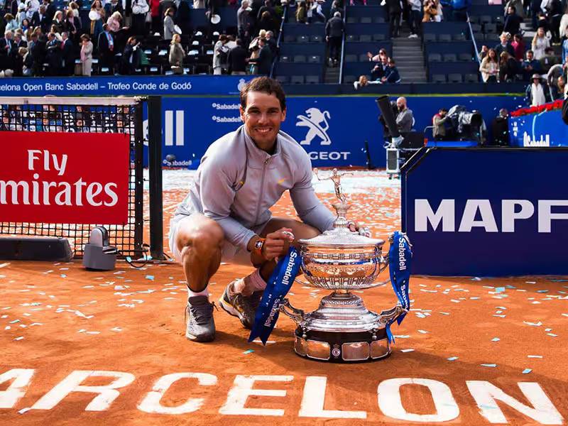 Rafael Nadal kneeling and smiling with the Barcelona Open trophy on a clay court, surrounded by tournament branding and spectators.