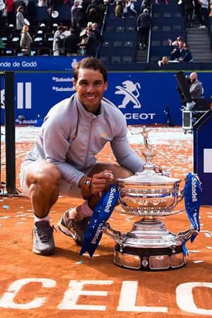 Rafael Nadal kneeling and smiling with the Barcelona Open trophy on a clay court, surrounded by tournament branding and spectators.