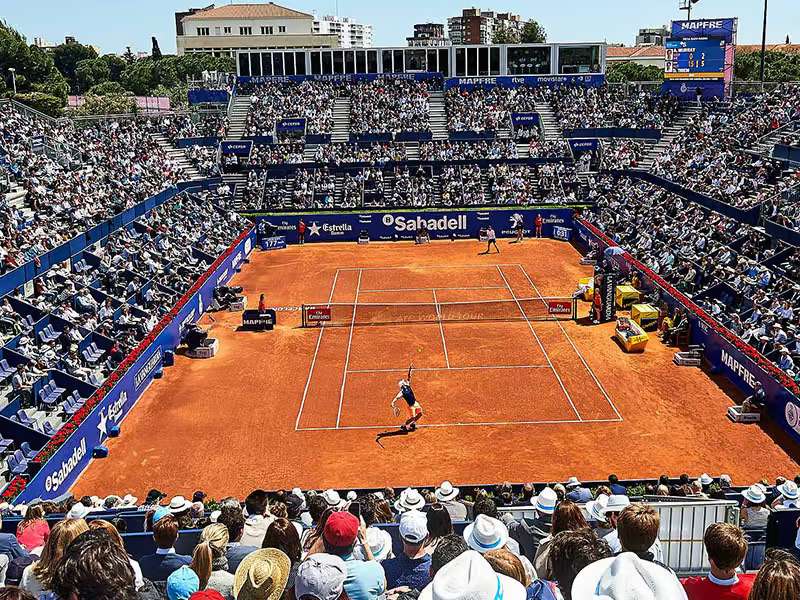 Wide shot of a professional tennis match on a clay court in Barcelona, with a packed stadium of spectators.