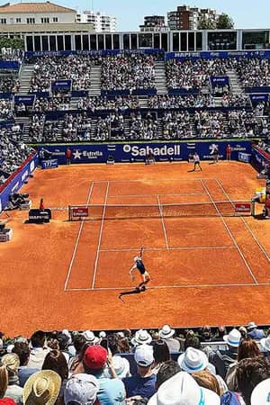 Wide shot of a professional tennis match on a clay court in Barcelona, with a packed stadium of spectators.