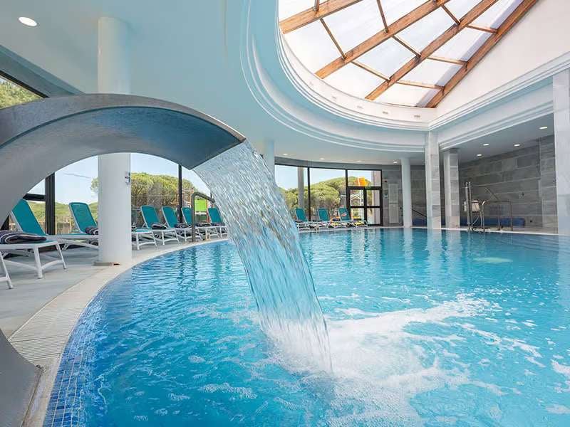 Indoor pool area at Barosa Park featuring a waterfall, sun loungers, and a bright skylight ceiling.